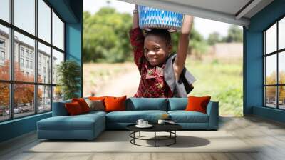 Grinning georgeous black African girl in a stylish dress balances a heavy bucket of water on her head Wall mural