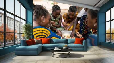 Group of coulorfully dressed black girls sitting around a big metal bowl, sharing a typical African meal made out of rice and vegetables Wall mural