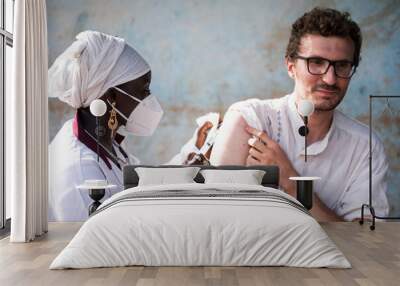 Young caucasian adult with a tense expression on his face receiving a vaccine shot in his right arm from a black nurse wearing a facial mask in an African hospital setting Wall mural