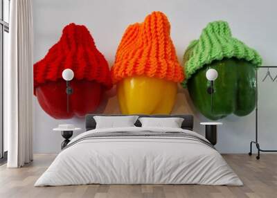 Raw, whole large red, orange and green peppers with the same colour loom knitted woollen hats lay on a white surface and background. Wall mural