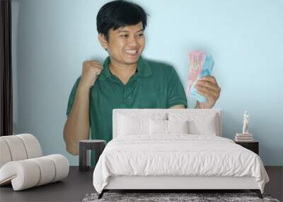 Young Asian man showing excited face expression with fist clenched while holding paper money. Wearing a green t-shirt Wall mural