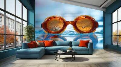 Orange swimming goggles floating in a pool, with water droplets on the lenses, evoking a sense of leisure and refreshment. Wall mural