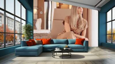 Confident beautiful positive young business lady in a luxurious brown overalls in a comfortable leather armchair in her hotel room. Charming woman enjoys apartments in a spa hotel during check-in Wall mural