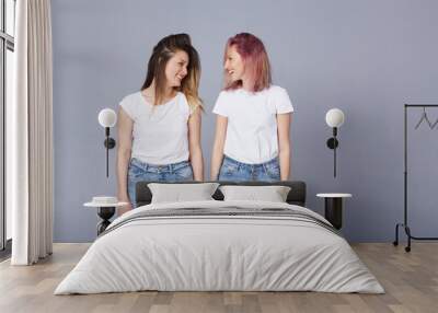 Horizontal portrait of two pretty women with happy expression, wears similar casual white t-shirt with denim, poses over gray background. Adorable lovely female student smile at each other. Copyspace Wall mural