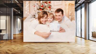 Merry Christmas and Happy Holidays! Cheerful parents and their cute daughter girls exchanging gifts. Parent and one little children having fun and playing together near Christmas tree indoors. Wall mural