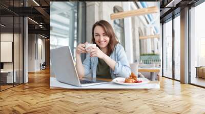 Portrait of beautiful young woman accountant or manager of large company sitting at table in cafe during lunch with mug of coffee and social networking using a laptop and wifi Wall mural