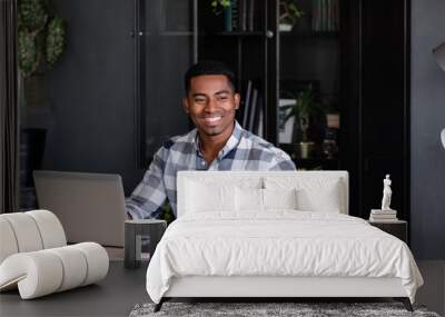 Positive young successful african american designer working on a new project while sitting at a table with a laptop. Handsome smiling mixed race businessman rejoices in career growth Wall mural