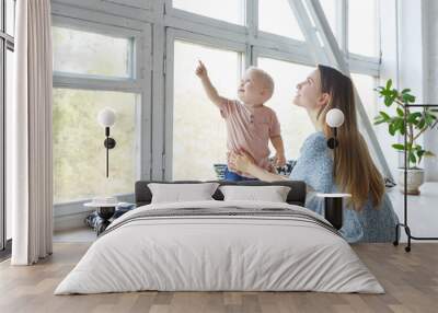 Side view of cheerful woman holding baby boy in her hands and looking at him with love sitting on wooden white floor over big window at home. Young beauty mom spending morning with two-year old son Wall mural