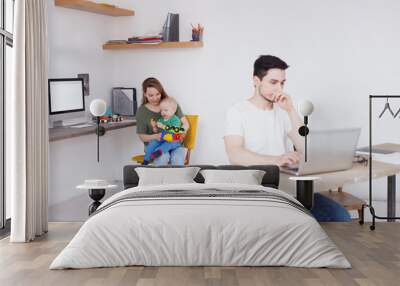 Stressed young father work all night at business project, siting in front of laptop, tries to concentrate on work at home. His wife and child in background. Overwork and sleep deprivation concept Wall mural