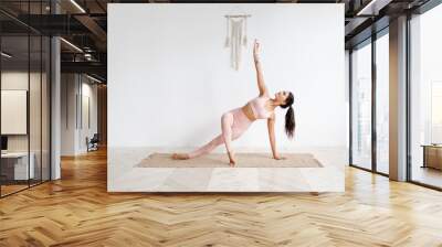 Young attractive woman practicing yoga in bright yoga class on wooden eco floors tanding in Full Version of Vasisthasana exercise, Side Plank pose, working out, pink sportswear. White wall background Wall mural