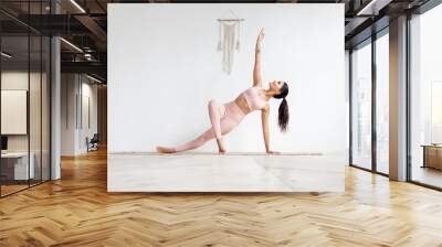 Young attractive woman practicing yoga in bright yoga class on wooden eco floors tanding in Full Version of Vasisthasana exercise, Side Plank pose, working out, pink sportswear. White wall background Wall mural