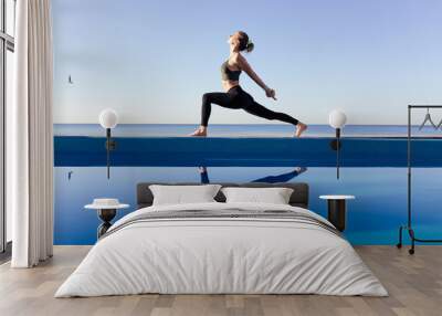 Young attractive woman practicing yoga stand in Warrior one exercise, Virabhadrasana pose, working out by the pool, above the beach, relaxing against blue sky. Health and beauty concept Wall mural