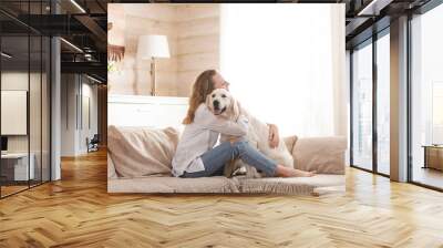 Young pretty woman in casual clothes hugging her beloved big white dog sitting on the sofa in the living room of her cozy country house. Animal communication concept Wall mural