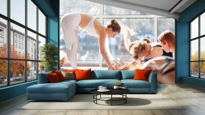 Young woman yoga instructor straightens her students while doing stretching while sitting on mats in the gym against the background of a large window. ?oncept of group lessons with a teacher Wall mural