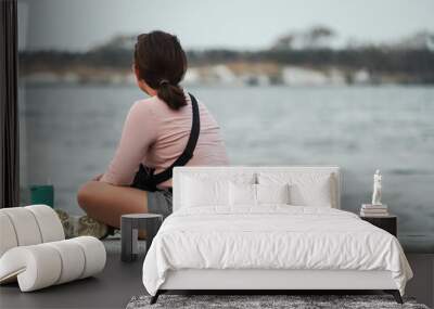 A young asian woman sits cross-legged, enjoying and contemplating the beachside view alone. Concept for mental health and loneliness Wall mural