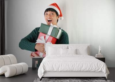 Excited young asian man wearing santa clause hat and green long sleeved sweater is carrying large gift box with ribbon, isolated over white background. Concept for Christmas Holiday and New Year Party Wall mural