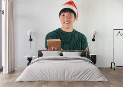 Excited young asian man wearing santa clause hat and green long sleeved sweater is carrying small gift box, isolated over white background. Concept for Christmas Holiday and New Year Party. Wall mural