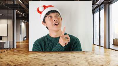 Excited young asian man wearing santa clause hat and green long sleeved sweater with smiling face expression, isolated over white background. Concept for Christmas Holiday and New Year Party Wall mural