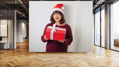 Excited young asian woman wearing santa clause hat and red long sleeved sweater is carrying large gift box with ribbon, isolated over white background. Concept for Christmas Holiday and New Year Party Wall mural