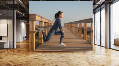 Sporty mid adult woman doing a leg stretch warm up exercise outdoors on a wooden boardwalk Wall mural