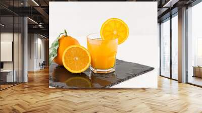 An inviting glass full of orange juice with orange slice on the rim, a half orange and a whole one with leaf on a square wet slate plate on white background Wall mural