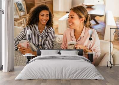 Elegance, beauty and happiness: multiracial young women couple having fun wearing pajama sitting on the sofa drink cup of tea. Afro american curly woman laughing at home with her best caucasian friend Wall mural