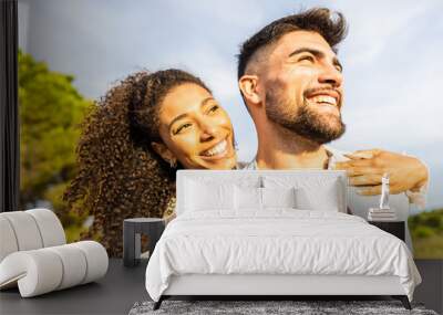 Multiracial happy young beautiful couple in love having fun outdoor in nature at dusk looking at horizon kissed by the setting sun. Toothy smiling Afro-American girl embracing from back her boyfriend Wall mural