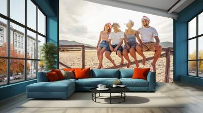 Suggestive photography with backlight sun effect of four young happy people enjoying outdoor life in exotic vacation resort sitting on wooden fence on a white sand beach at sunset Wall mural