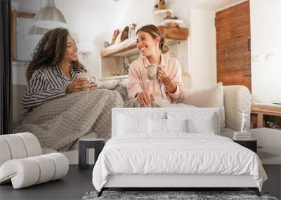 Two fine young women sitting on the sofa with a blanket on their legs laughing enjoying the house in winter having tea with pastries - Mixed race female couple home lifestyle Wall mural