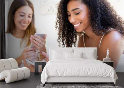 Two young beautiful women at home using smartphone - Cute brunette black Hispanic having fun indoor chatting on social networks with her beautiful blonde female friend - Focus on curly woman Wall mural