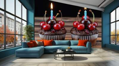 Two chocolate cupcakes topped with cherries and lit candles, arranged on a wooden table.

 Wall mural