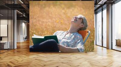 Senior woman reading book and laughing with head back in sunny field Wall mural