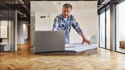 Senior male architect using laptop by blueprint at desk in incomplete house Wall mural