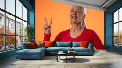 Portrait of mature woman giving peace sign hand gesture, isolated on orange background in studio. People emotions, lifestyle concept Wall mural