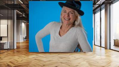 Portrait of stylish mature woman wearing black hat smiling and laughing, isolated on blue background with copy space Wall mural