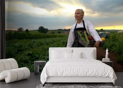 Wide angle view of stylish, smiling, female chef carrying a crate of vegetables on her hip and smelling fresh picked corn. Wall mural