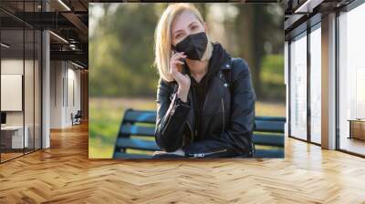 Woman using a Smart phone in the park sitting on a bench in a park on a sunny day, wearing black medical mask for protection, during Covid-19 quarantine. Wall mural