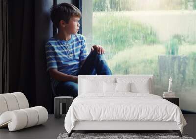 Little boy, home and watching the rain by window of natural water drops and thinking in relax indoors. Child relaxing by windowsill looking out glass at storm in lonely or sad for weather at house Wall mural