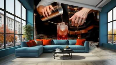 Bartender pouring a cocktail into a glass with ice at a bar counter, focus on hands and shaker. Wall mural