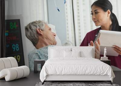 Asian woman doctor talking with senior Caucasian male patient lying in hospital bed. Medical professional monitoring symptoms of ill patient Wall mural