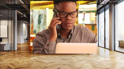 Black man talks on a phone at a restaurant while working on his laptop Wall mural