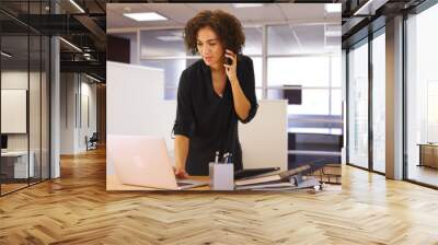 Businesswoman using laptop computer in office while listening on smartphone device Wall mural