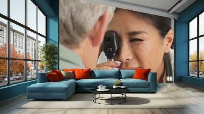 Close up on doctor using ophthalmoscope to look into eyes of senior male patient. Japanese woman primary care physician or ophthalmologist doing check up Wall mural