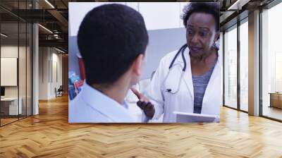 Close view of black medical doctor discussing health issues with young male patient. Senior African doctor using portable tablet to take notes of patients health Wall mural