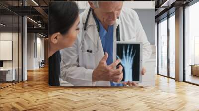 Doctor using tablet computer with x-ray of hand to talk about recovery with female patient. Senior Caucasian male medical professional with screen demonstrating arthritis to woman Wall mural