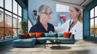 Female doctor using stethoscope listening to senior patients heart. Older woman visiting her primary care physician for a regular check up Wall mural