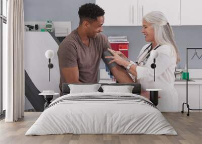 Handsome african-american patient seated in medical room having his blood pressure checked by senior doctor. Mature white female physician checking male patients blood pressure in health clinic Wall mural