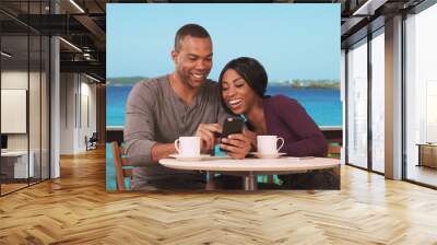 Happy Black couple on honeymoon sitting at restaurant with ocean view drinking coffee and using smartphone. African man and woman drinking coffee on vacation using phone to book next trip Wall mural