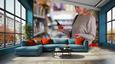 Happy senior woman looking at grocery store aisle with cellphone in hand Wall mural