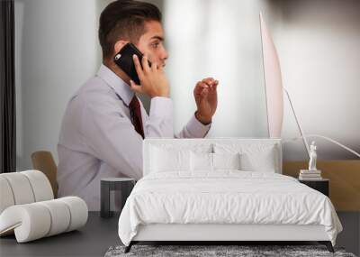 Hispanic business man working on computer while talking on cellphone. Mexican businessman making phone call and using pc. Young Millennial Latino office worker in his 20s Wall mural